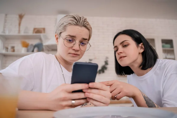 Молодая лесбийская пара смотрит на смартфон на кухне — стоковое фото