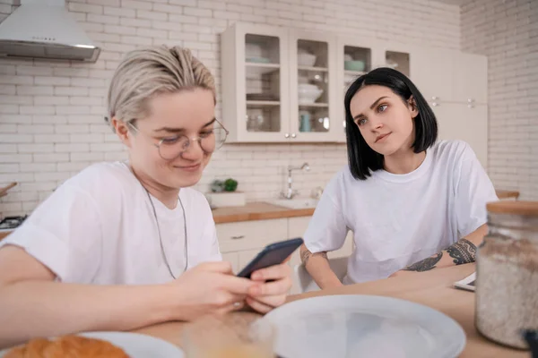 Татуированная женщина смотрит на подругу с помощью смартфона на кухне — стоковое фото