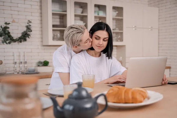 Mujer joven besar novia mientras se utiliza el ordenador portátil en la cocina - foto de stock