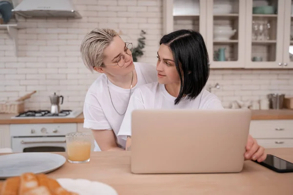 Mujer joven mirando a su novia usando el ordenador portátil en la cocina - foto de stock