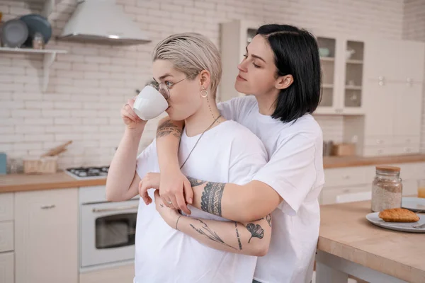 Brunette woman hugging tattooed girlfriend drinking coffee in kitchen — Stock Photo