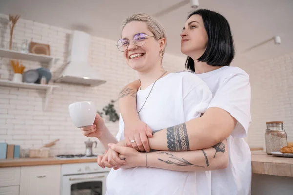 Brunette woman hugging tattooed girlfriend with cup of coffee in kitchen — Stock Photo