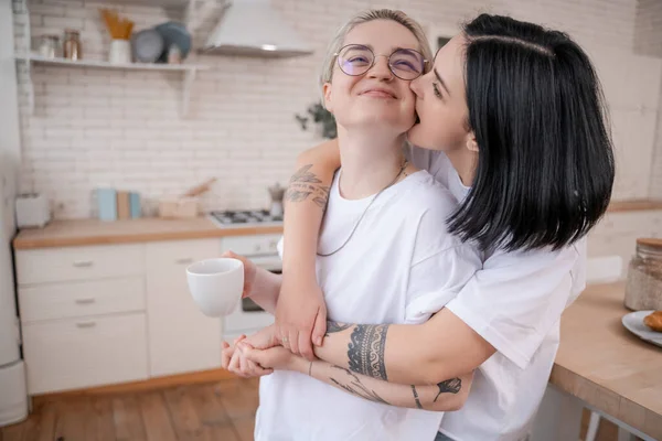 Femme brune mordant joue de petite amie tatouée avec tasse de café dans la cuisine — Photo de stock