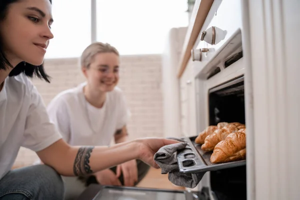 Tattooed lesbian couple looking at oven tray with freshly baked croissants in kitchen — Stock Photo