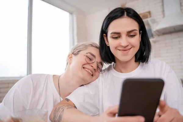 Morena mujer usando smartphone cerca feliz novia en cocina - foto de stock