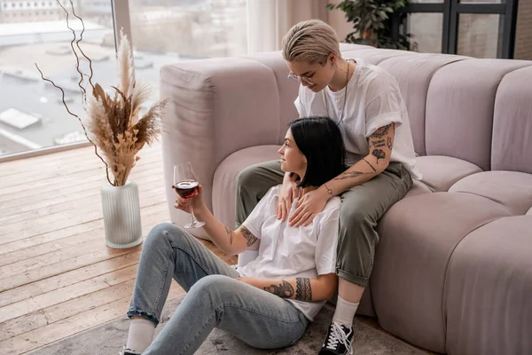 Femme tatouée regardant petite amie heureuse avec un verre de vin rouge — Photo de stock