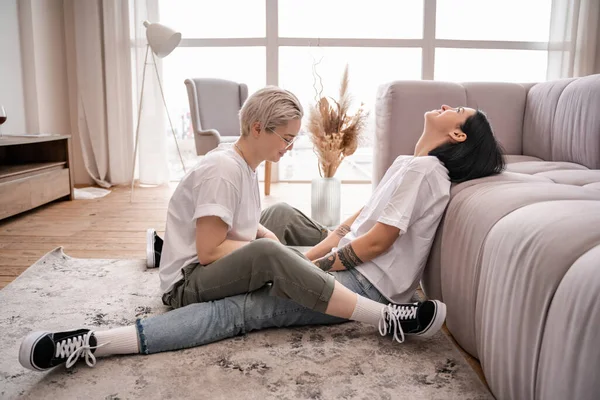 Tätowierte Frau sitzt mit brünetter Freundin auf Teppich in der Nähe von Couch — Stockfoto