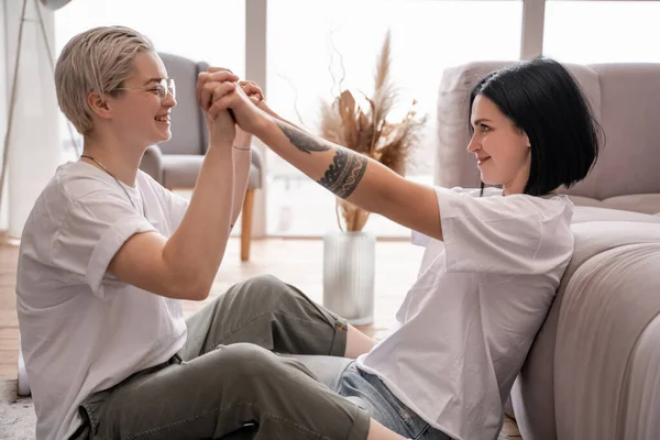 Vue latérale du couple lesbien heureux tenant la main dans le salon — Photo de stock