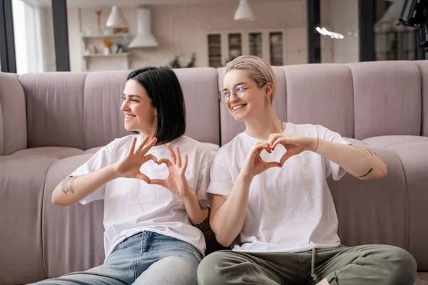 Sonriente lesbiana pareja mostrando corazón signo con las manos en sala de estar - foto de stock