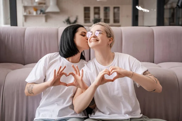 Feliz casal lésbico mostrando sinal de coração com as mãos na sala de estar — Fotografia de Stock