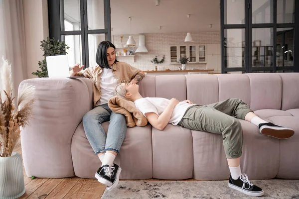 Morena mujer sentada en el sofá cerca de la computadora portátil y mirando a la novia - foto de stock