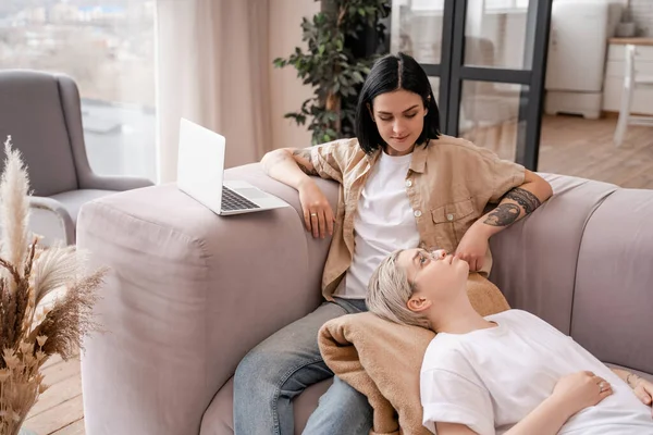 Tattooed woman sitting on couch near laptop and looking at girlfriend — Stock Photo