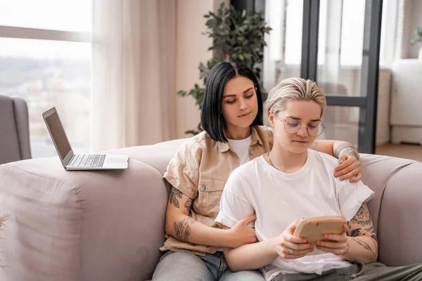 Lesbiana jugando en mbira mientras está sentada en el sofá con su novia cerca del ordenador portátil - foto de stock