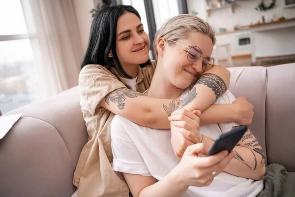 Pareja de lesbianas abrazándose mientras están sentados en el sofá y usando el teléfono inteligente - foto de stock