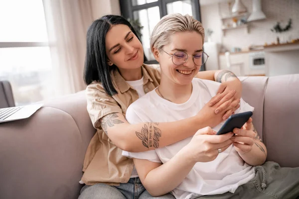 Feliz pareja lesbiana mirando el teléfono inteligente mientras se enfría en el sofá - foto de stock