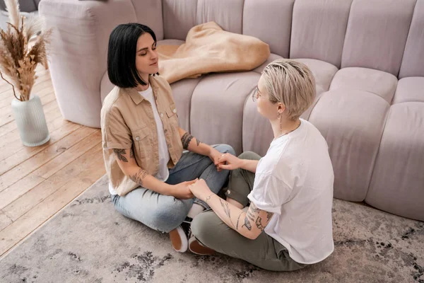 Pareja de lesbianas sentados en la alfombra y tomados de la mano en la sala de estar - foto de stock