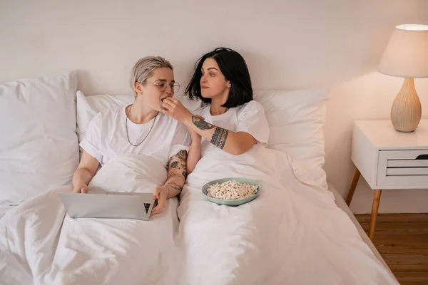 Brunette woman feeding girlfriend with popcorn near laptop on bed — Stock Photo