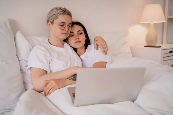 Junges lesbisches Paar umarmt sich, während es auf dem Bett liegt und sich Film auf Laptop ansieht — Stockfoto