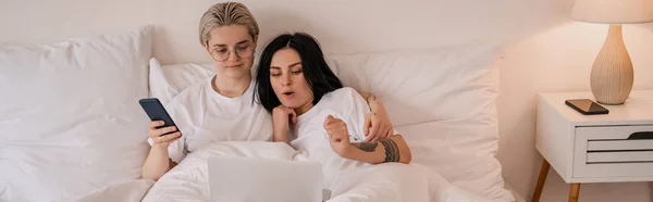 Молодая лесбийская пара лежит в постели и смотрит фильм на ноутбуке, баннер — стоковое фото