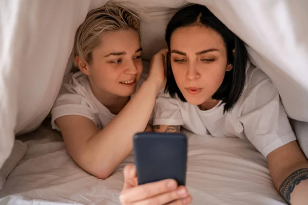 Junge Frau schreibt SMS auf Smartphone, während sie mit Freundin unter Decke liegt — Stockfoto