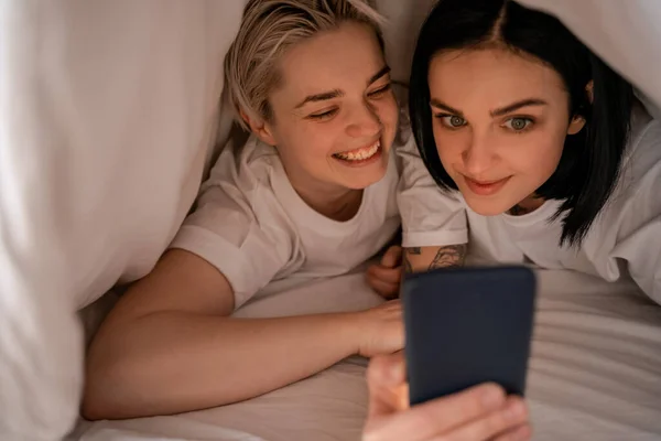 Junge Frau schreibt Nachrichten auf Smartphone, während sie mit Freundin unter Decke liegt — Stock Photo