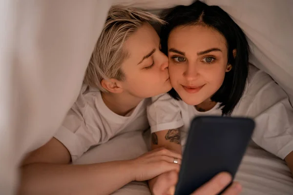 Jeune femme couché sous couverture et embrasser petite amie textos sur smartphone — Photo de stock