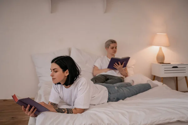 Brunette woman lying on bed and reading book near girlfriend on blurred background — Stock Photo