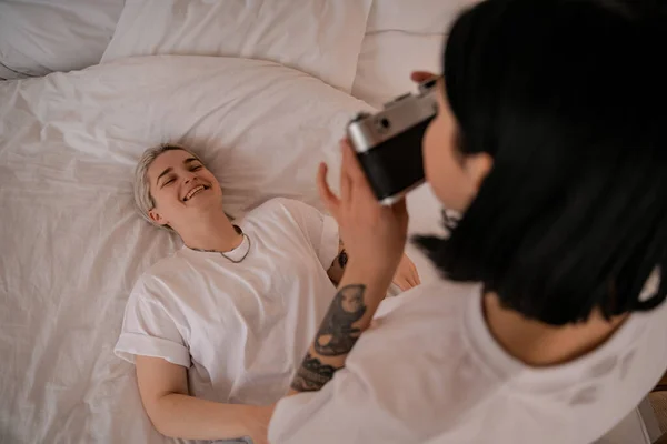 Blurred woman taking photo of girlfriend lying on bed and smiling on retro camera — Stock Photo