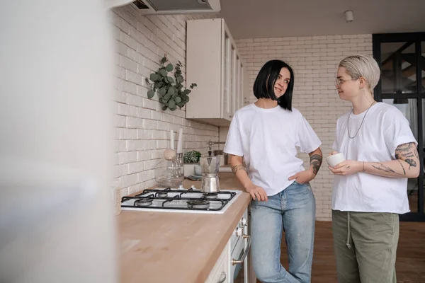 Junge Frau hält Tasse in der Hand und schaut Freundin in der Nähe von Kaffeekanne auf Herd an — Stockfoto