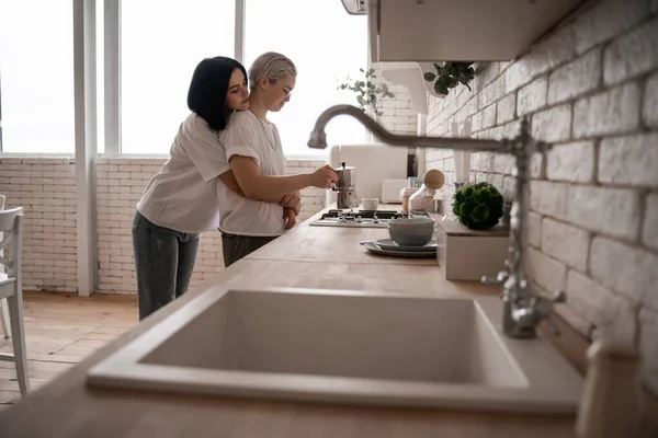 Giovane donna abbracciando fidanzata vicino caffettiera sul fornello con lavandino in primo piano sfocato — Foto stock