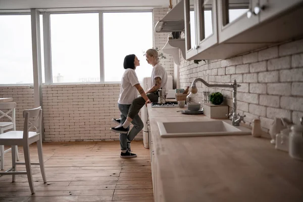 Sonriente joven sentada en la mesa de la cocina y mirando a la novia cerca de la cafetera en la estufa - foto de stock