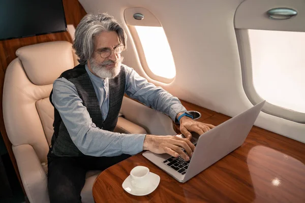 Зрелый бизнесмен, использующий ноутбук возле кофе в самолете — стоковое фото