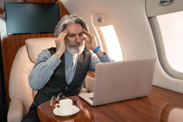 Уставший бизнесмен смотрит на ноутбук рядом с кофе и очки в реактивном самолете — стоковое фото