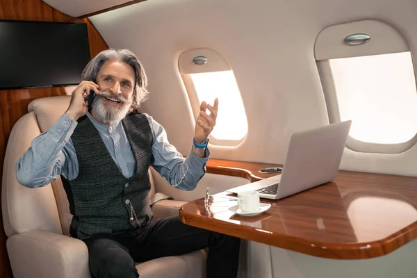 Uomo d'affari sorridente che parla su smartphone vicino a laptop e caffè in aereo — Foto stock