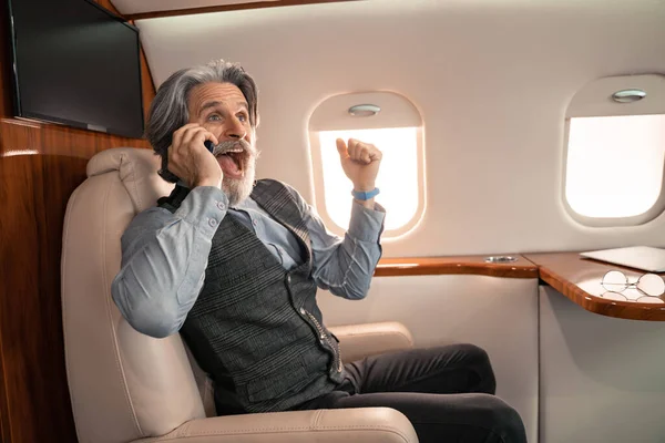 Eccitato uomo d'affari che parla su smartphone vicino agli occhiali in aereo privato — Foto stock