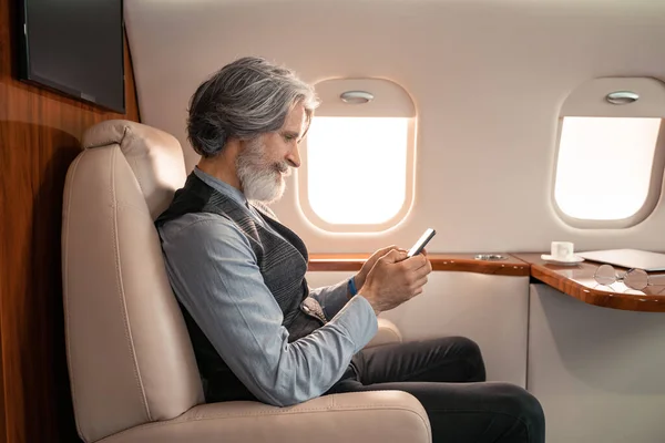 Vista lateral de un hombre de negocios maduro usando un teléfono inteligente cerca de anteojos y taza borrosa en jet - foto de stock