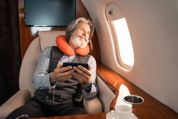 Uomo d'affari sorridente nel cuscino del collo che tiene lo smartphone mentre guarda la finestra dell'aereo — Foto stock