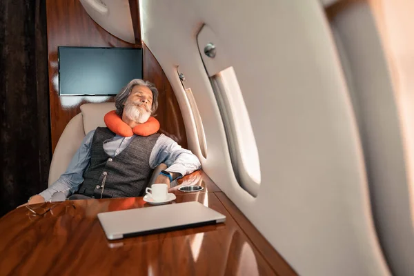 Mature businessman in neck pillow sleeping near laptop and coffee in plane — Stock Photo