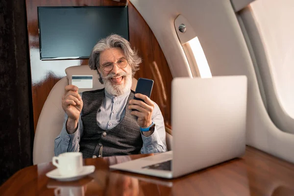 Homme d'affaires avec carte de crédit et smartphone clin d'oeil près d'un ordinateur portable et café en jet — Photo de stock