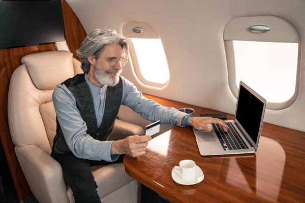 Mature businessman using credit card and laptop near coffee in private jet — Stock Photo