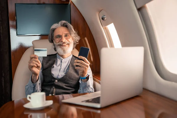 Усміхнений бізнесмен тримає кредитну картку і смартфон біля ноутбука і кави в приватному літаку — стокове фото
