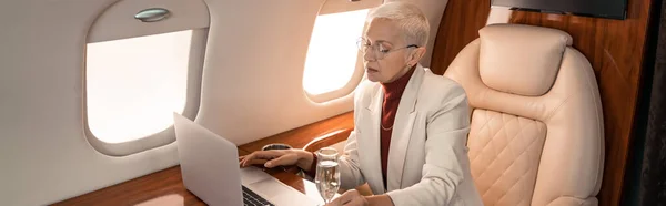 Femme d'affaires utilisant un ordinateur portable près d'un verre de champagne en jet, bannière — Photo de stock