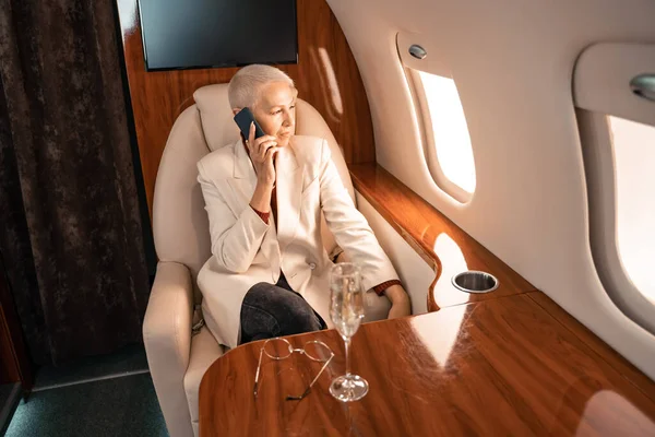 Доросла бізнесменка розмовляє на смартфоні біля розмитого шампанського і окулярів у літаку. — стокове фото