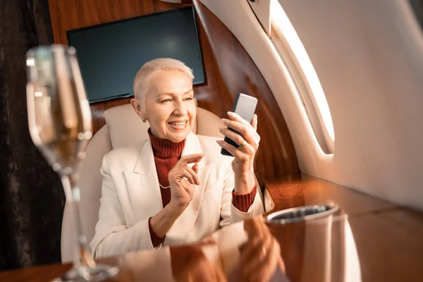 Smiling businesswoman using smartphone near blurred glass of champagne in plane — Stock Photo