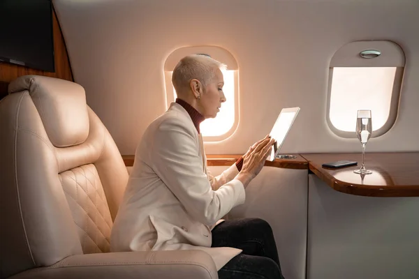 Side view of mature businesswoman using digital tablet near smartphone and glass of champagne in plane — Stock Photo