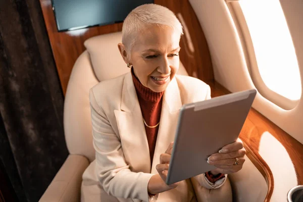 Mature businesswoman smiling while using blurred digital tablet in private jet — Stock Photo