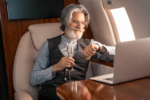 Улыбающаяся деловая женщина с шампанским смотрит на наручные часы возле размытого ноутбука в самолете — стоковое фото
