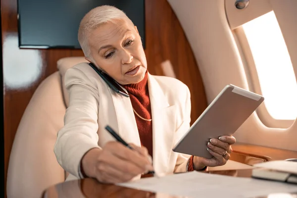 Geschäftsfrau mit digitalem Tablet schreibt auf Papier und spricht im Privatflugzeug auf dem Smartphone — Stockfoto