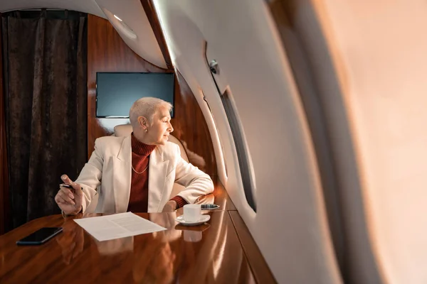 Sonriente mujer de negocios madura mirando la ventana del avión cerca del café y el papel - foto de stock