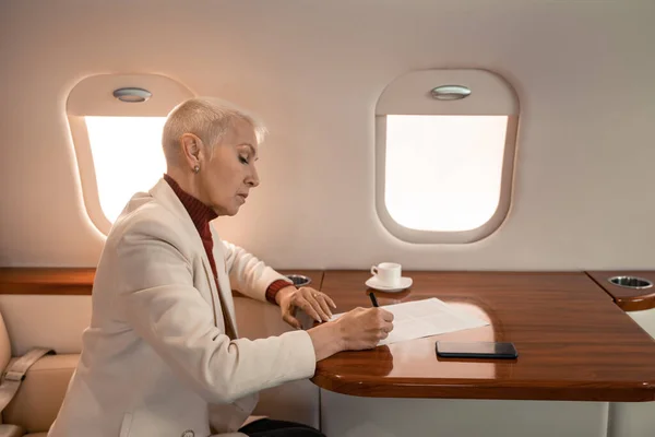 Vista lateral de la mujer de negocios escribiendo en papel cerca de café y teléfono inteligente en jet privado - foto de stock